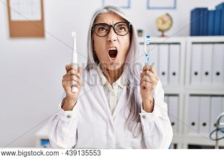 Middle age grey-haired woman working at dentist clinic holding electric teethbrush and toothbrush angry and mad screaming frustrated and furious, shouting with anger looking up. 