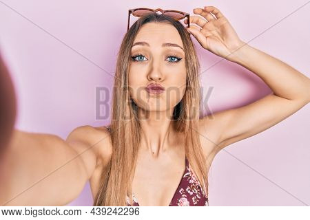 Young blonde girl wearing bikini taking a selfie puffing cheeks with funny face. mouth inflated with air, catching air. 