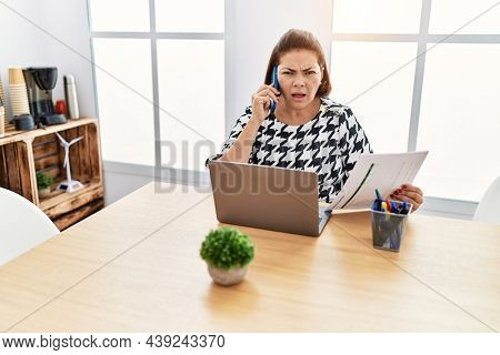 Middle age hispanic woman working at the office with laptop speaking on the phone in shock face, looking skeptical and sarcastic, surprised with open mouth 
