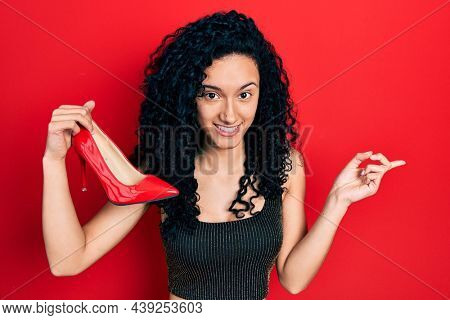 Young hispanic woman with curly hair holding red high heel shoes smiling happy pointing with hand and finger to the side 
