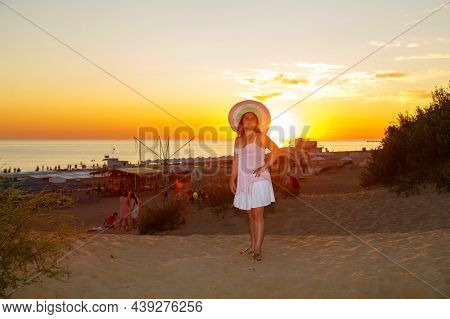 Woman In A White Dress And A Straw Hat On A Background Of The Sea At Sunset. Horizontal Photo