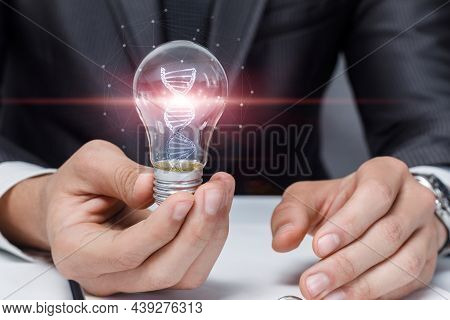 Business Man Shows A Light Bulb With Glowing Dna Molecules On A Blurred Background.