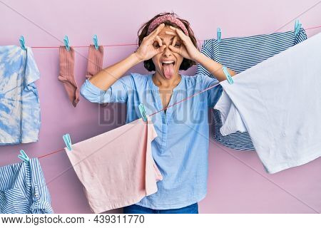 Young brunette woman doing laundry around string hangs doing ok gesture like binoculars sticking tongue out, eyes looking through fingers. crazy expression. 