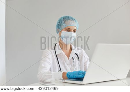 Indoor Shot Of Concentrated Woman Doctor Therapist Wearing Gown, Surgical Mask, Medical Cap And Glov