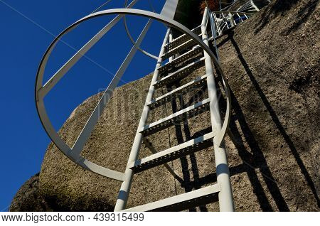 Galvanized Iron Gray Fire Ladder Leading To The Rock. Safety Is Ensured By A Tube-shaped Grille Of T