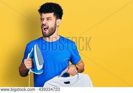 Young arab man with beard holding electric steam iron and white t shirt angry and mad screaming frustrated and furious, shouting with anger. rage and aggressive concept. 
