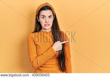 Young brunette teenager wearing casual yellow sweater pointing aside worried and nervous with forefinger, concerned and surprised expression 