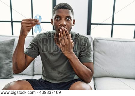 Young african american man holding blue ribbon sitting on the sofa at home covering mouth with hand, shocked and afraid for mistake. surprised expression 