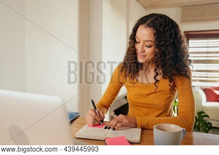 Happy Multi-cultural Female Focussed, Writing In Notebook. Young Professional Remote Working With La