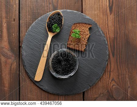 Black Paddlefish Caviar In A Wooden Spoon, A Slice Of Bread And A Jar On A Black Round Graphite Boar