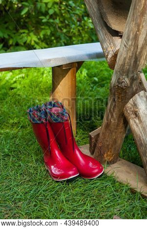 Vertical Photo Of Red Rubber Boots Near A Wooden Table And A Bench On Green Grass In A Village In Su
