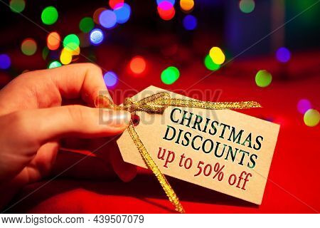 A Woman's Hand Holds A Label With Text - Christmas Discounts Up To 50 Off. On A Red Background, With