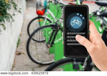 technology, transport and sustainability concept - close up of hand holding smartphone with mobile app and bicycle icon on screen over electric bike parking and charging station in city on background