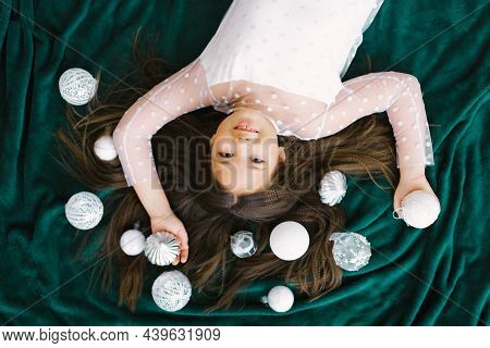 Cute Girl With Long Hair With Christmas Decor, White Christmas Toys Lying On An Emerald Blanket