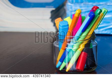 Multi-colored Pens And Markers In A Stand On The Background Of Stack Of Notebooks.  Pack Of Blue Sch