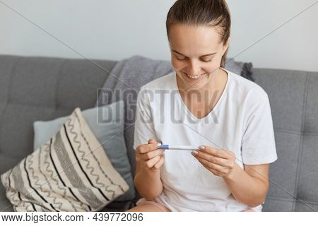 Indoor Shot Of Happy Young Adult Woman Wearing White T Shirt Holding Pregnancy Test In Hands, Waitin
