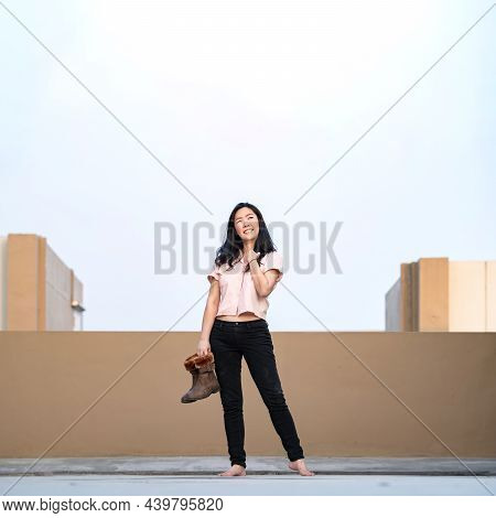 Asian Woman Stands And Smiles Alone With Holds Boots On Rooftop In Pastel Blue - Brown Shade.