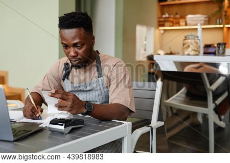 Young African American Man Wearing Apron Sitting At Table In Front Of Laptop In Modern Small Cafe Ta