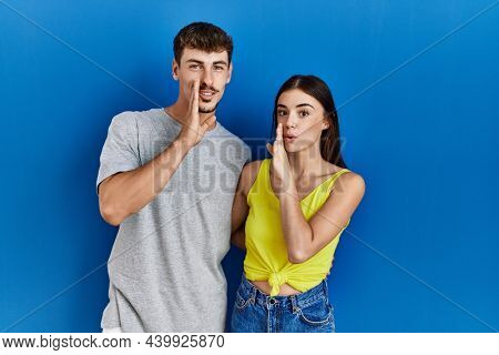 Young hispanic couple standing together over blue background hand on mouth telling secret rumor, whispering malicious talk conversation 