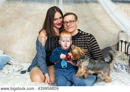 A Little Redhead Boy Sits On A Bed Next To Perents And A Dog. Happy Family With Dog. Christmas Time