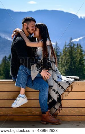 A Loving Couple Wrapped In A Blanket Sitting On The Veranda Of A Mountain Villa On The Background Of