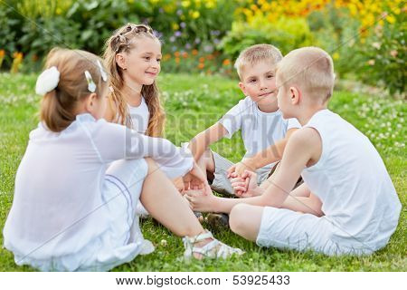 Four children sit on grass at lawn holding hands, one boy yalks, other listen and look at him