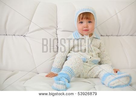 Cute little kid in knitted suit and hat sits on white couch at home. Shallow depth of field.