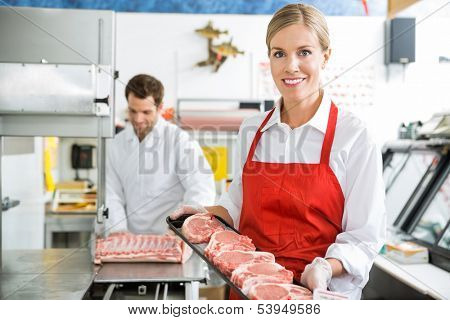 Portrait of beautiful butcher holding meat tray at store with colleague working in background