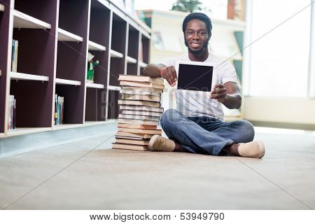 Full length portrait of smiling young student with stacked books showing digital tablet while sitting on floor at library