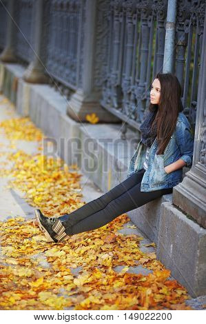 Young pensive brunette girl sitting on a concrete curb around a beautiful iron fence under the feet of yellow autumn maple leaves.