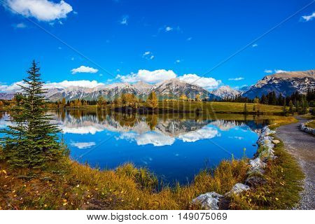 Path around the lake. Canmore, near Banff National Park. Majestic mountains and scenic cumulus clouds are reflected in the water. The concept of recreational tourism