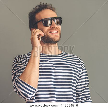 Handsome stylish young man in sailor's striped vest and sun glasses is talking on the mobile phone and smiling on gray background