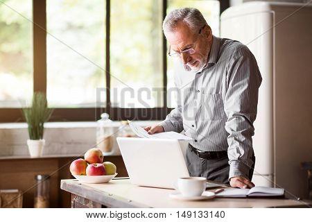 Business in the morning. Good-looking grey enterpreneur in glasses working with laptop and papers while having coffee and breakfast