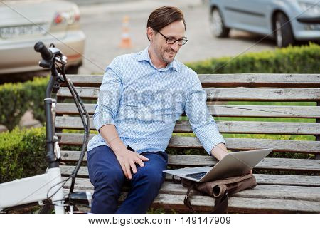 What a wonderful work. Glad handsome bespectacled man working on laptop and smiling while sitting on the bench.