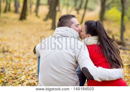 Beautiful young couple in autumn park sitting on the ground covered with yellow leaves, kissing. Back view.