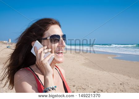 portrait of brunette woman looking side with sunglasses smiling and talking on mobile phone smartphone at beach with sea and blue sky behind