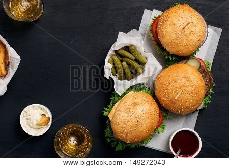 Three different burger on white paper on dark surface with pickles ketchup mayonnaise fries and light beer top view with copy space