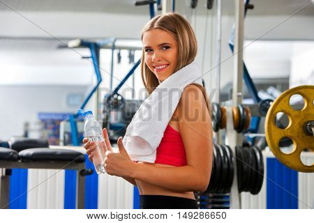 Woman with towel working his arms and chest at gym. Girl relax near barbell. Sport girl keeps bottle water and thumb up.