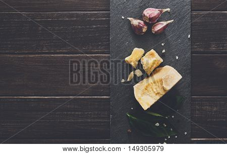 Cheese delikatessen closeup on black stone desk at wooden surface background. Parmesan pieces decorated with basil and garlic, top view image with copy space