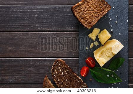 Cheese delikatessen closeup and healthy rye bread on black stone desk at wood background. Parmesan pieces decorated with basil and cherry tomatoes, top view image with copy space, soft toning