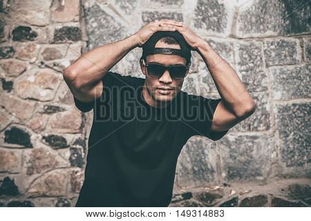 Young and cool. Handsome young African man in casual clothes holding hands on cap and looking at camera while standing against the stoned wall outdoors