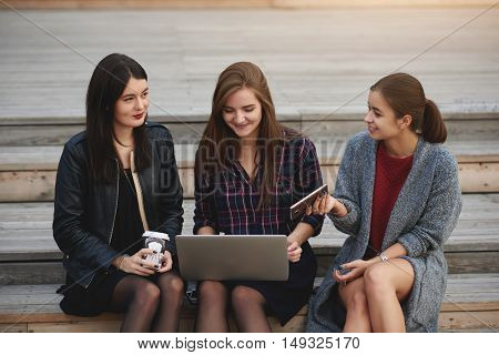 Hipster girls with trendy look are using net-book for on-line booking cinema tickets while are sitting outdoors. Woman is searching in network via net-book movie to watch while her friends talking