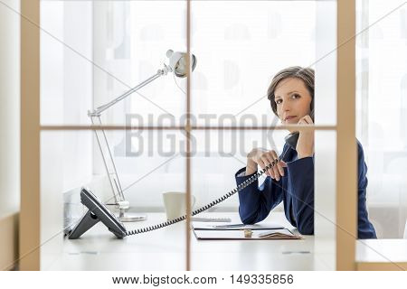 Businesswoman or manageress talking on a phone in the office glancing at the camera through a glass partition with a serious expression.