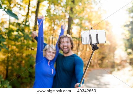Beautiful young runners in colorful sunny autumn forest taking selfie with smartphone on selfie stick