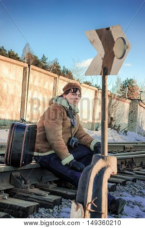 Serious young man in a sheepskin coat and beret with a suitcase and a camera sitting on the rails near the sign of railway switch in winter day.