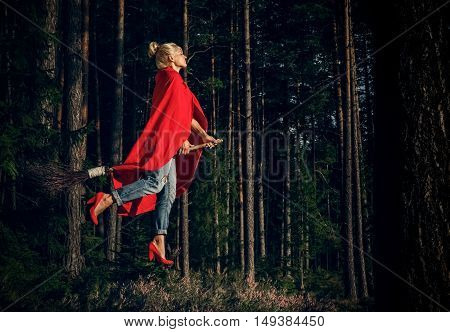 Modern witch blond in jeans and a red cape flying on a broom through the forest. Levitation. Halloween holiday.