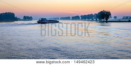 Dutch river with a freigther and trees along the banks just before sunrise in the beginning of the fall season.
