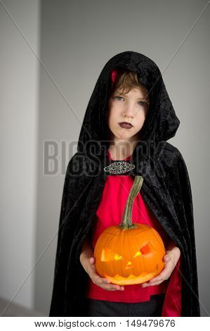 All Hallows Eve. Boy age dressed in a costume for Halloween. He represents the evil wizard. Boy is gowned in a black-red toga with a hood. He holds pumpkin with a candle inside - Halloween symbol.