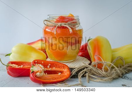 Bell pepper preserved in a glass jar with fresh peppers on white wooden background. Homemade marinated in oil red pepper. Glass jar with conserved roasted sweet paprika. Selective focus.