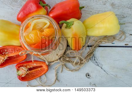 Bell pepper preserved in a glass jar with fresh peppers on white wooden background. Homemade marinated in oil red pepper. Glass jar with conserved roasted yellow and red paprika. Top view. Copy space.